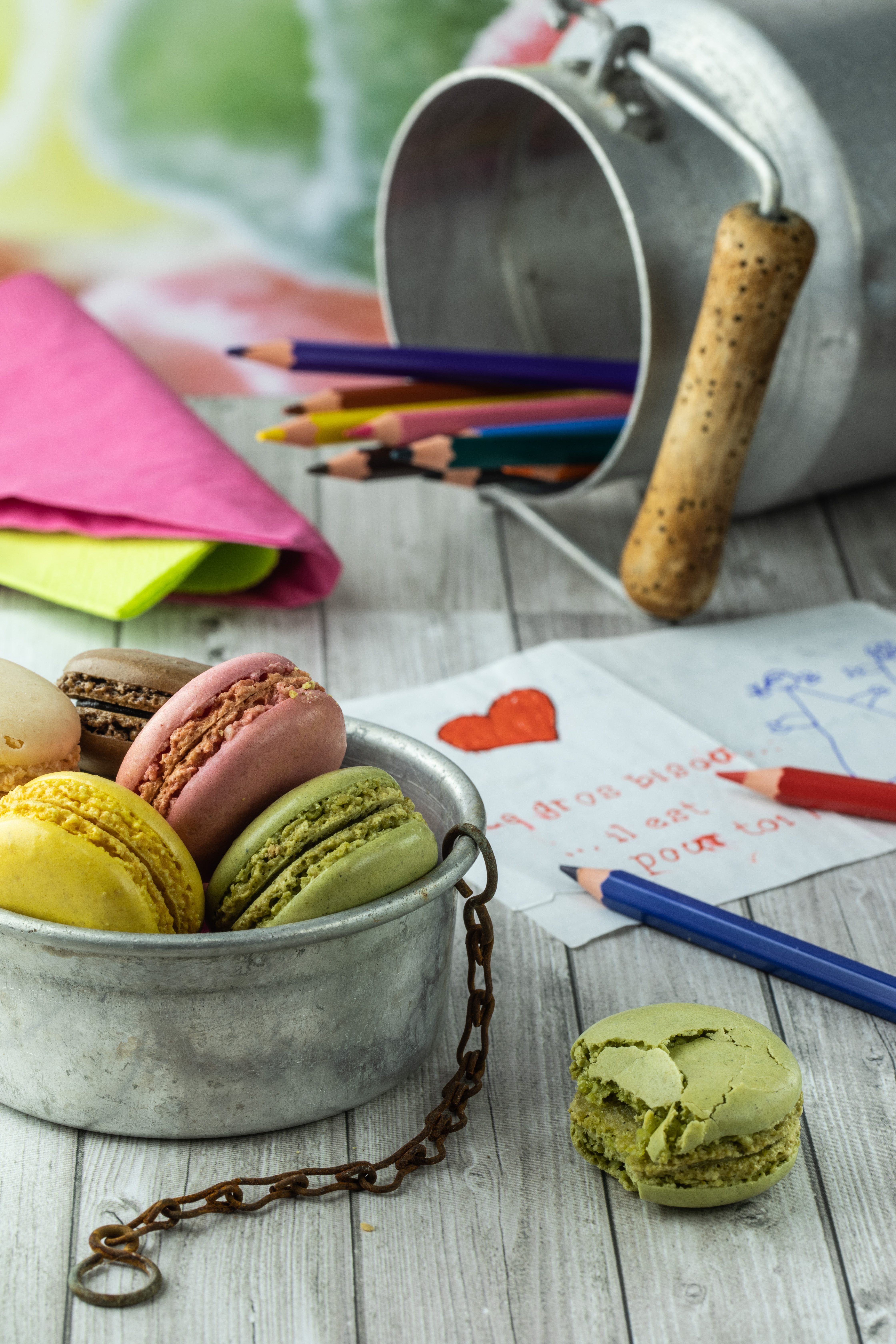 Photo de divers objets sur une table : une coupelle en métal avec des macarons de différentes couleurs, un pot à lait en métal avec des crayons de couleur à l'intérieur, un dessin enfantin