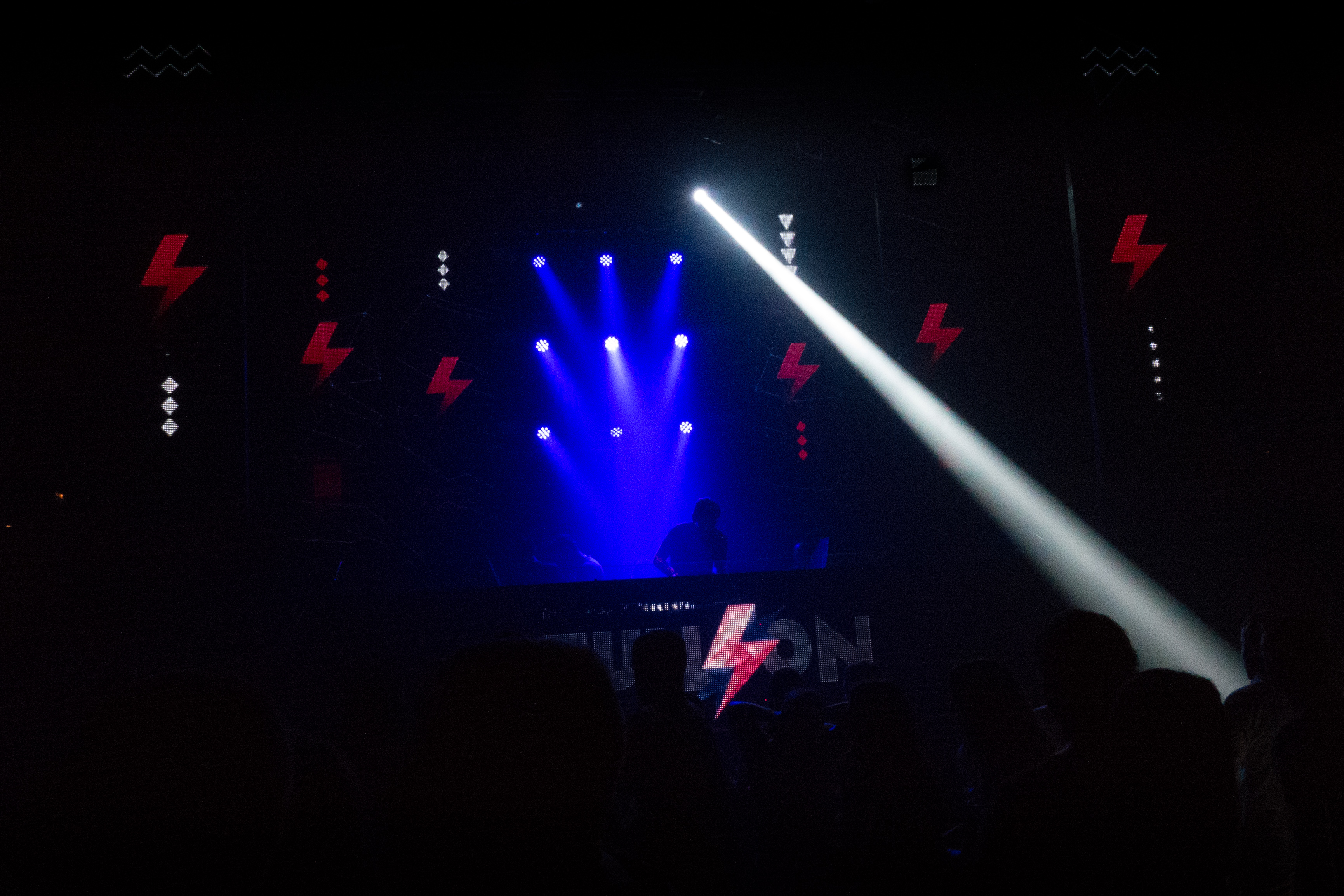 Photo in a very dark nightclub, with white and blue spotlights, red lights in the shape of lightning, and a DJ silhouette at the decks
