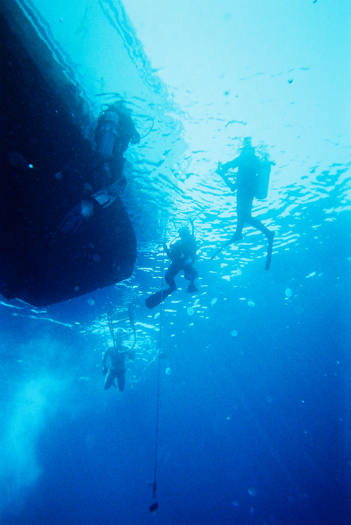 Underwater photo taken from the bottom to the surface of the water, with a boat hull and 4 divers